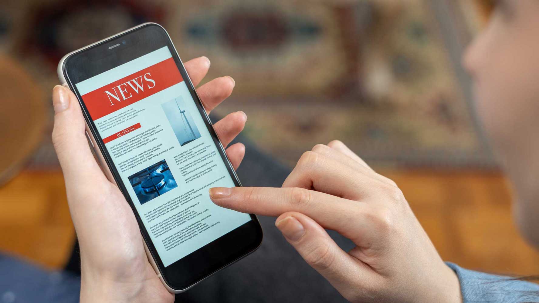 A close-up shot of a person's hand scrolling through a news article titled "NEWS" on a smartphone, representing the modern access to information and media literacy.