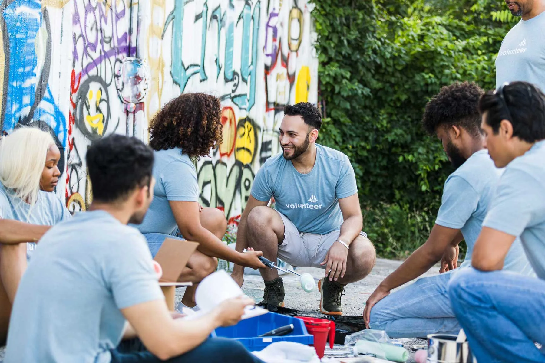 A diverse group of young adult volunteers sitting and kneeling together, discussing their community mural painting project in front of a graffiti-covered wall.