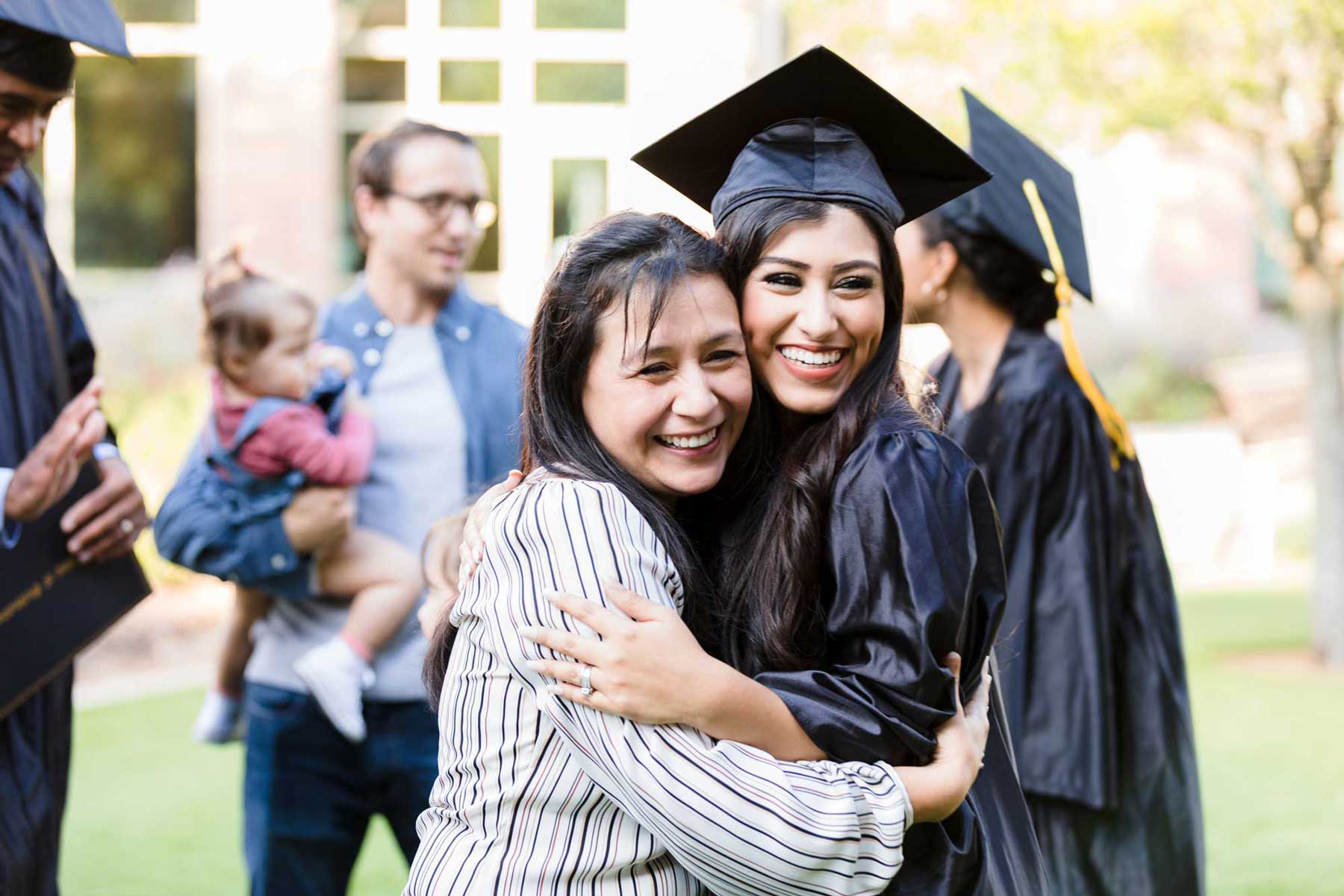 A smiling young woman in a graduation cap and gown tightly hugs her proud mother during an outdoor commencement ceremony.