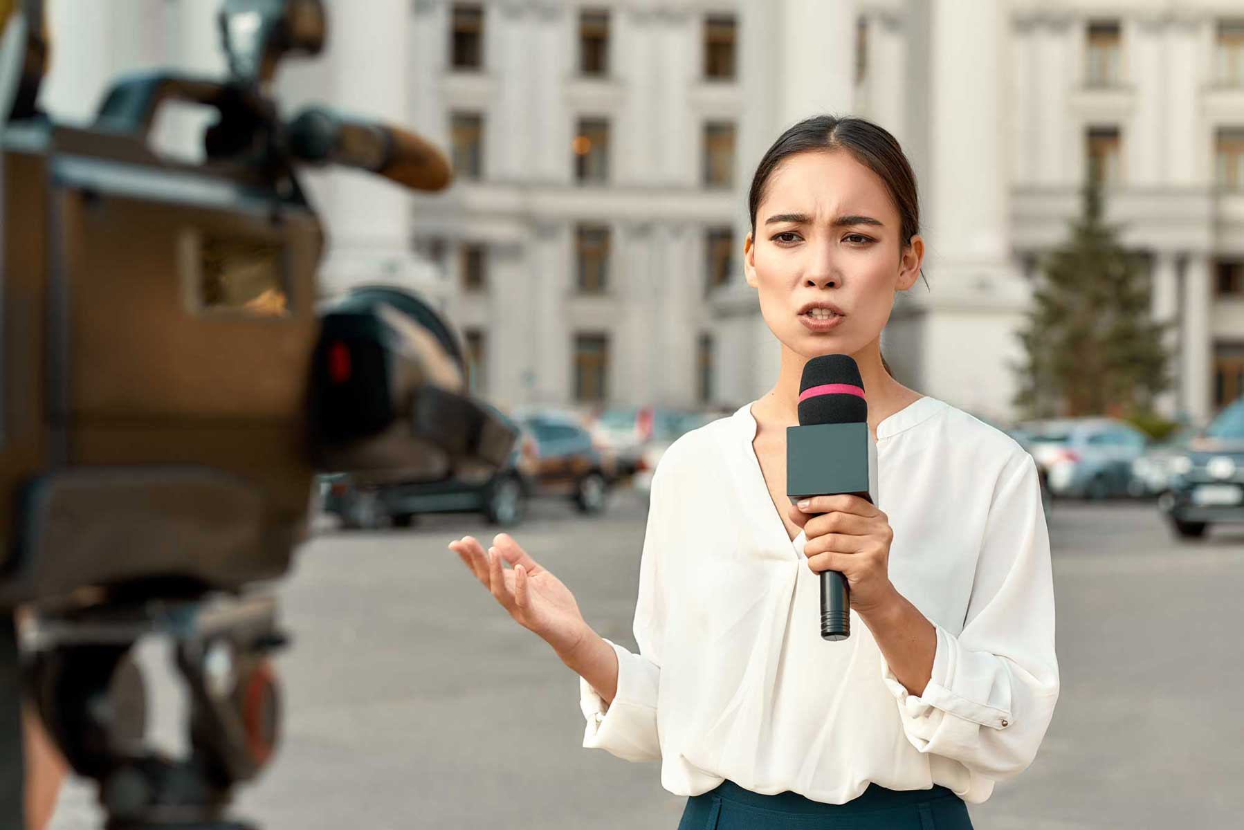 A female news reporter holds a microphone and speaks seriously into a camera, standing in front of a government building, representing the importance of a free press.
