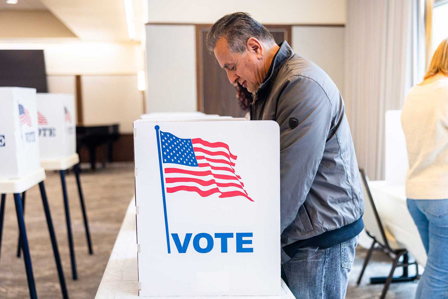 A man leaning over a voting booth with a graphic of the US flag and the word "VOTE" clearly visible, representing the fundamental right of civic participation in an election.