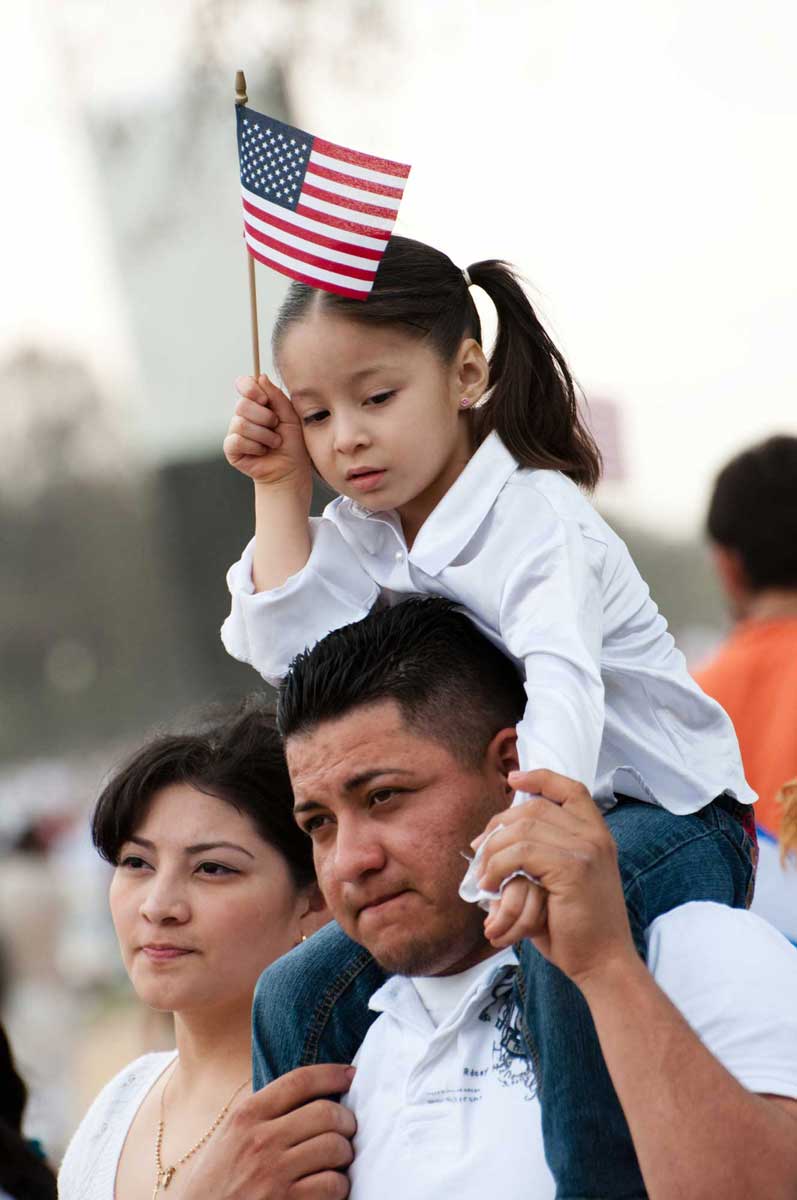 A Hispanic father carries his young daughter on his shoulders as she holds a small American flag, standing next to the mother at an outdoor public gathering.