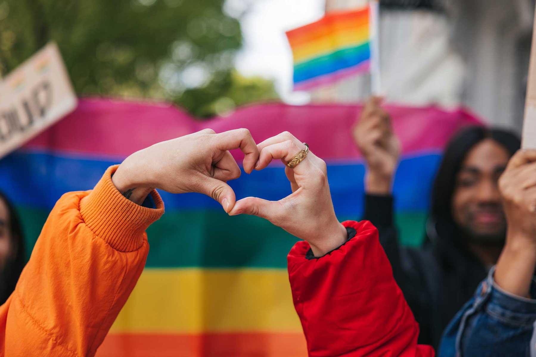 A close-up of two people making a heart shape with their hands in front of a blurred rainbow flag, representing the fight for equality and inclusive democratic values.