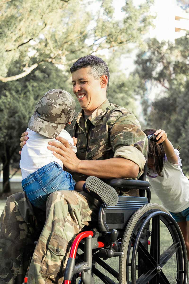 A smiling soldier in a wheelchair wearing camouflage fatigues hugs a young child on his lap while another child pushes the wheelchair from behind in a park setting.