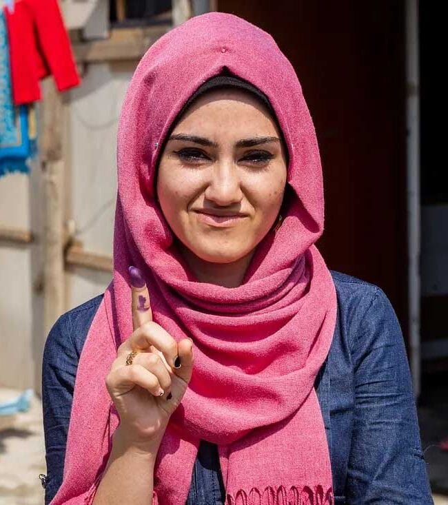 A smiling young Iraqi woman wearing a pink hijab holds up her finger to show the purple ink mark, indicating she has successfully voted in an election.