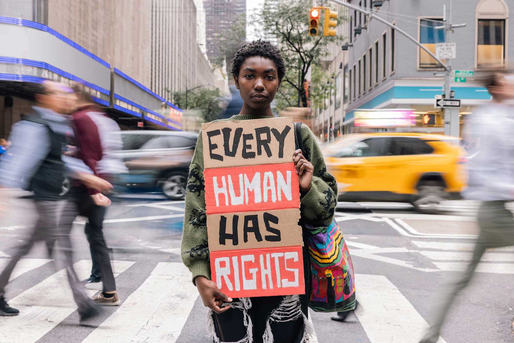 A determined young Black person standing in a busy city street holding a handmade sign that reads, "EVERY HUMAN HAS RIGHTS," advocating for universal human rights.